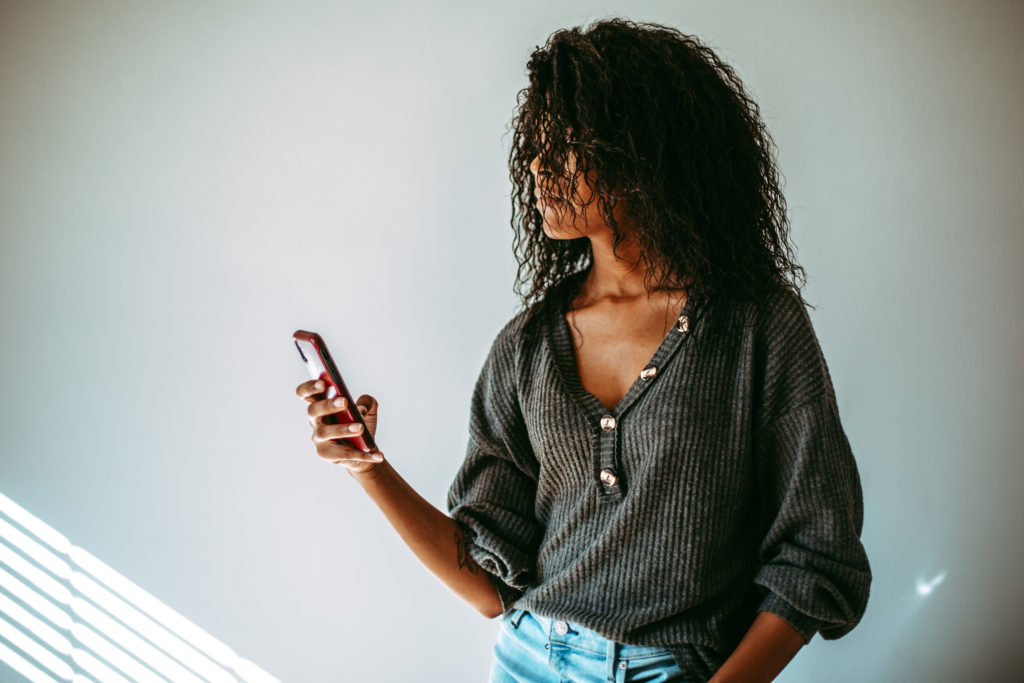 African American girl looking down at her cell phone, wearing a grey shirt with puffy sleeves and light blue high waisted jeans. 