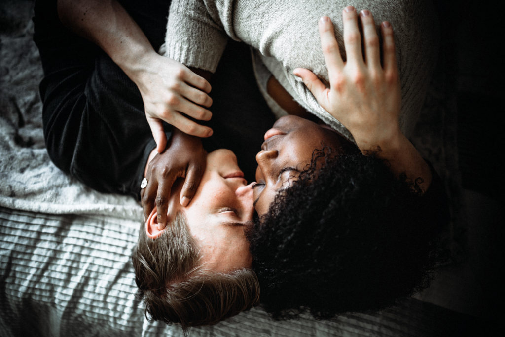 An African American female and Caucasian male embracing each other on a bed. Their eyes are closed and they are lightly smiling. 