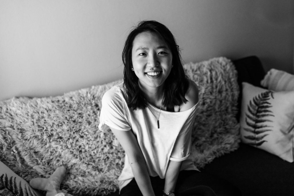 An Asian woman around her 20's, smiling at the camera while sitting on her couch. She has a comfortable white off the shoulder tee. 