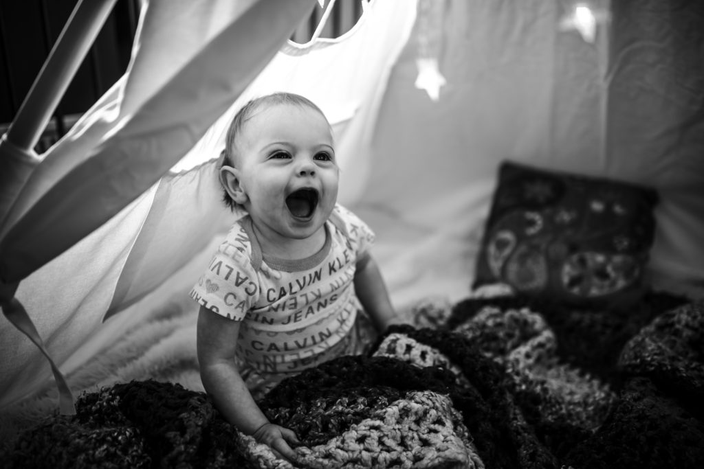 A one year old girl inside a tent, laughing at something in the distance.