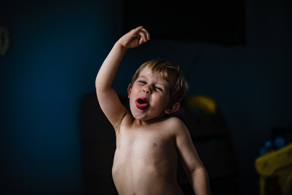 A photo of a little boy in front of a dark background, He is lit by the sunlight and sticking his tongue out, dancing.