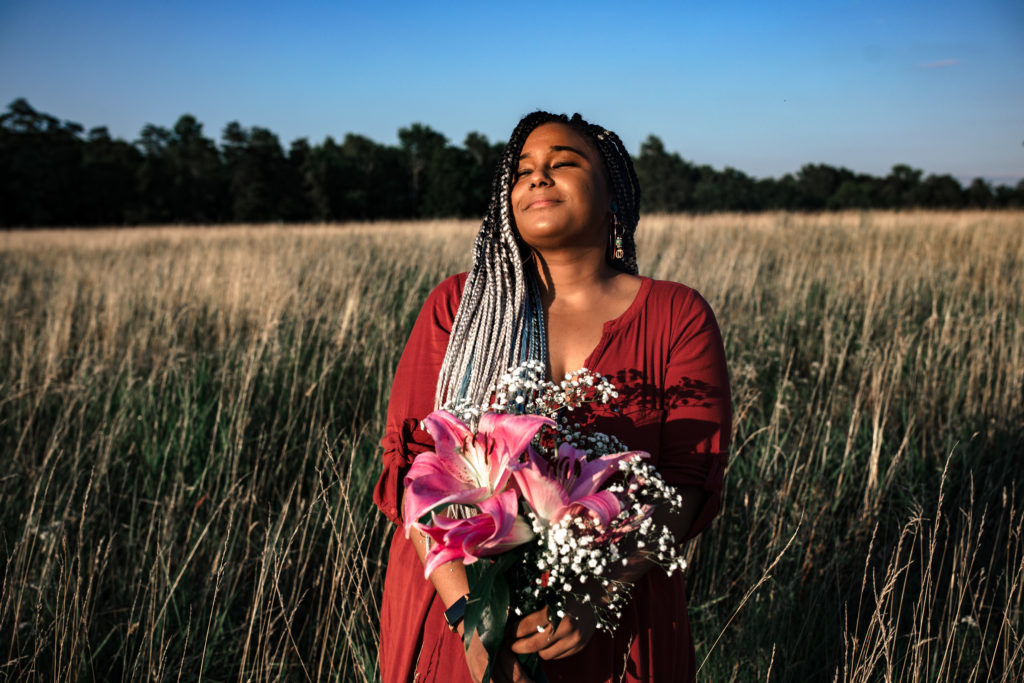 black female with ombre black and white braids and a red dress. Holding stargazer flowers. She is standing in a field of tall grass.
