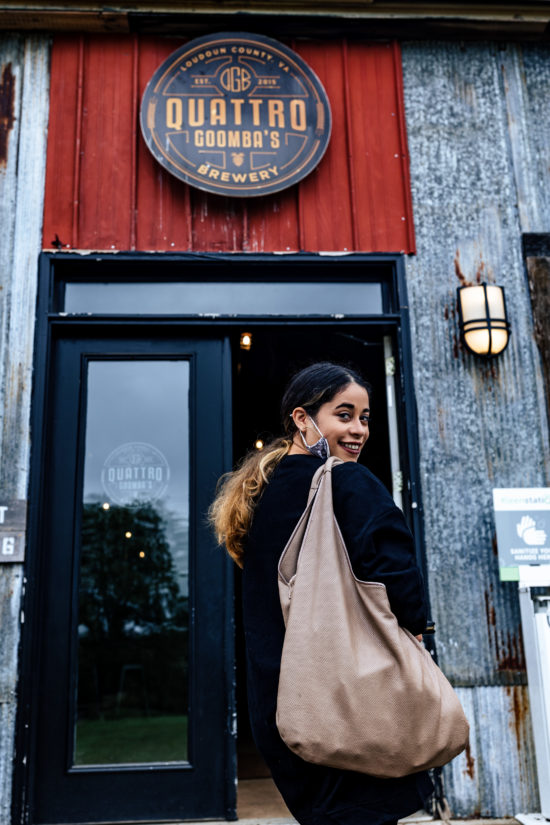 Female smiling and standing in front of a brewery called Quattro Goombas. She has a brown bag and black sweatshirt on with long black and blond ombre hair. 