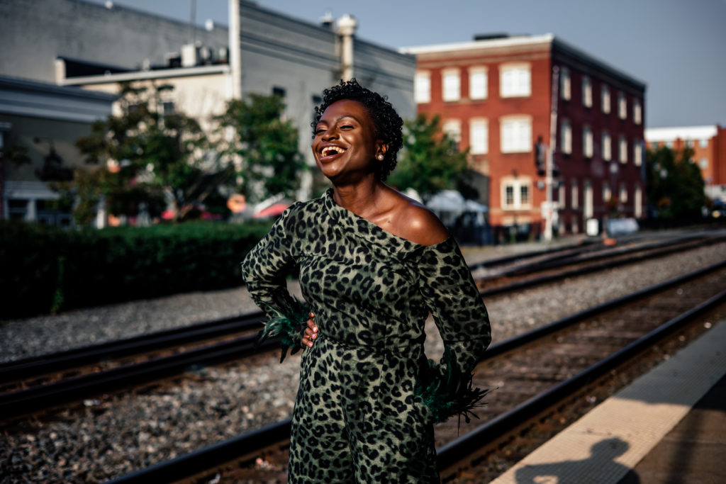 An African American woman is standing on a train platform in front of train tracks. She is wearing a green cheetah print jumper. She is laughing. 