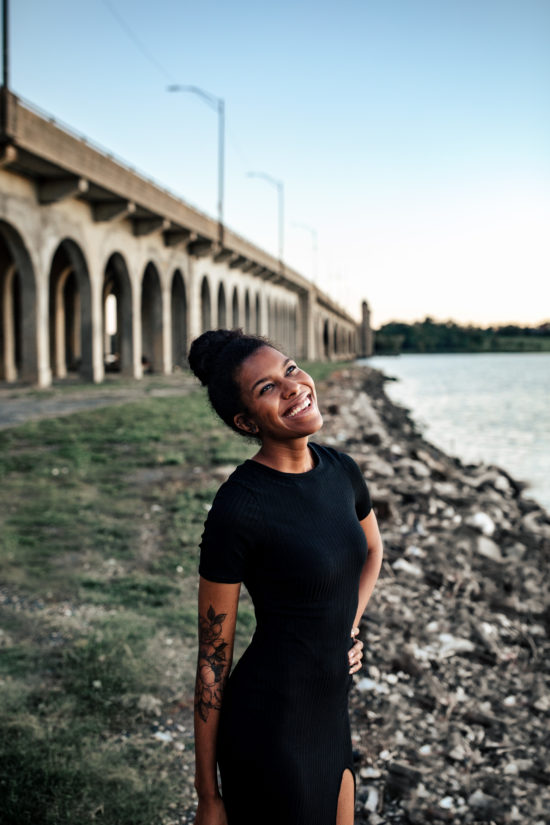 An African American Woman wearing a form fitting black dress is smiling at the sky. She has a flower tattoo on her arm. She is standing in front of a bridge and water.