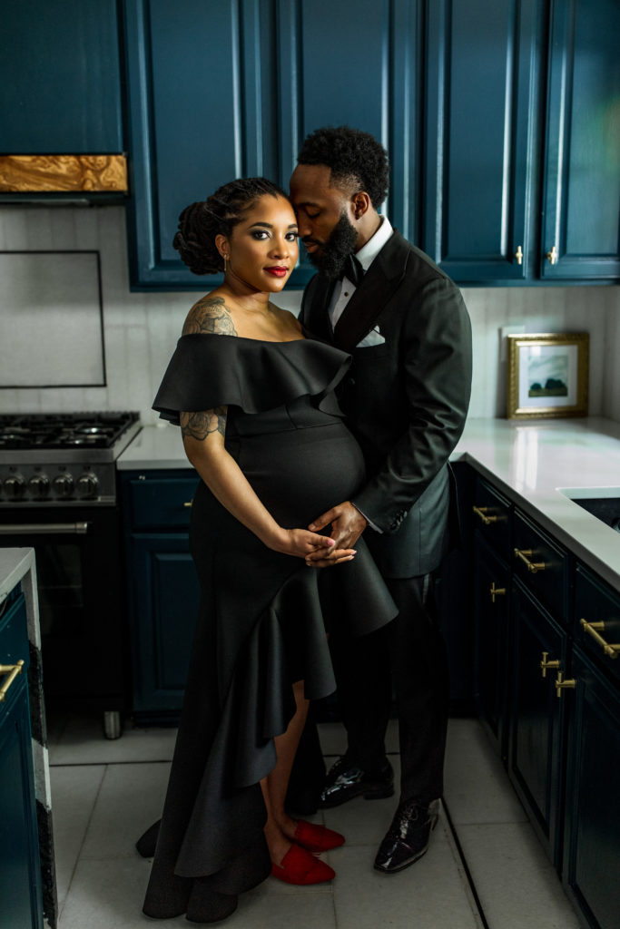 In home maternity session with Bewitched Photography. A black couple is dressed in in a black suit and dress for their couples photo session in an eclectic kitchen.