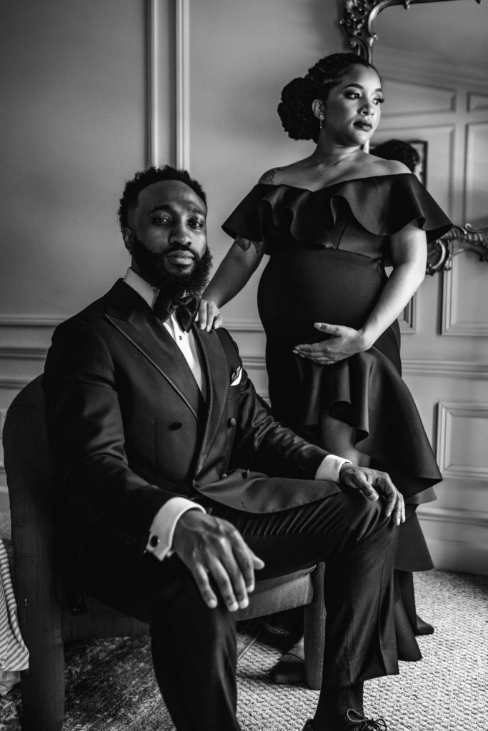 maternity session with Bewitched Photography. A black couple is dressed up in in a black suit and dress for their couples photo session in an eclectic bedroom.