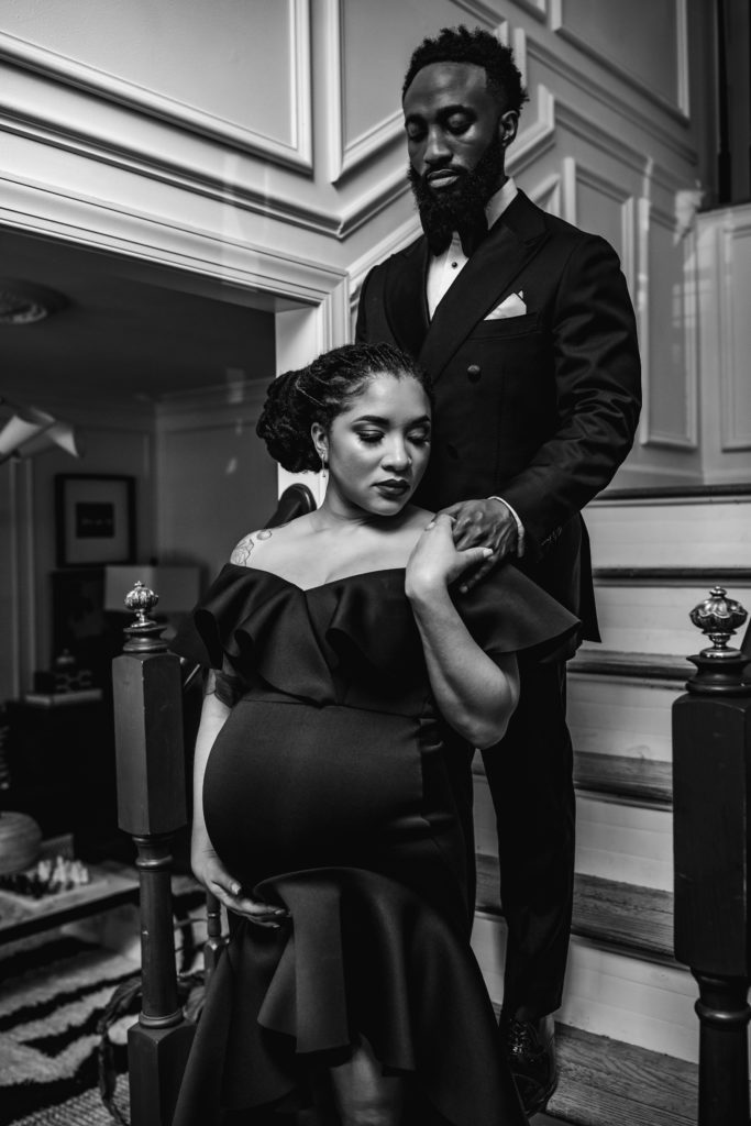 In home maternity session with Bewitched Photography. A black couple is dressed up in in a black suit and dress for their couples photo session.